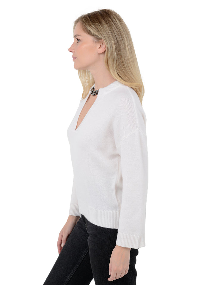 Side view of model wearing Audrina in cloud white. The sweater is relaxed and made of 100% cashmere. The sweater also features a v-neck, drop shoulder, and silver chain trim across neckline.