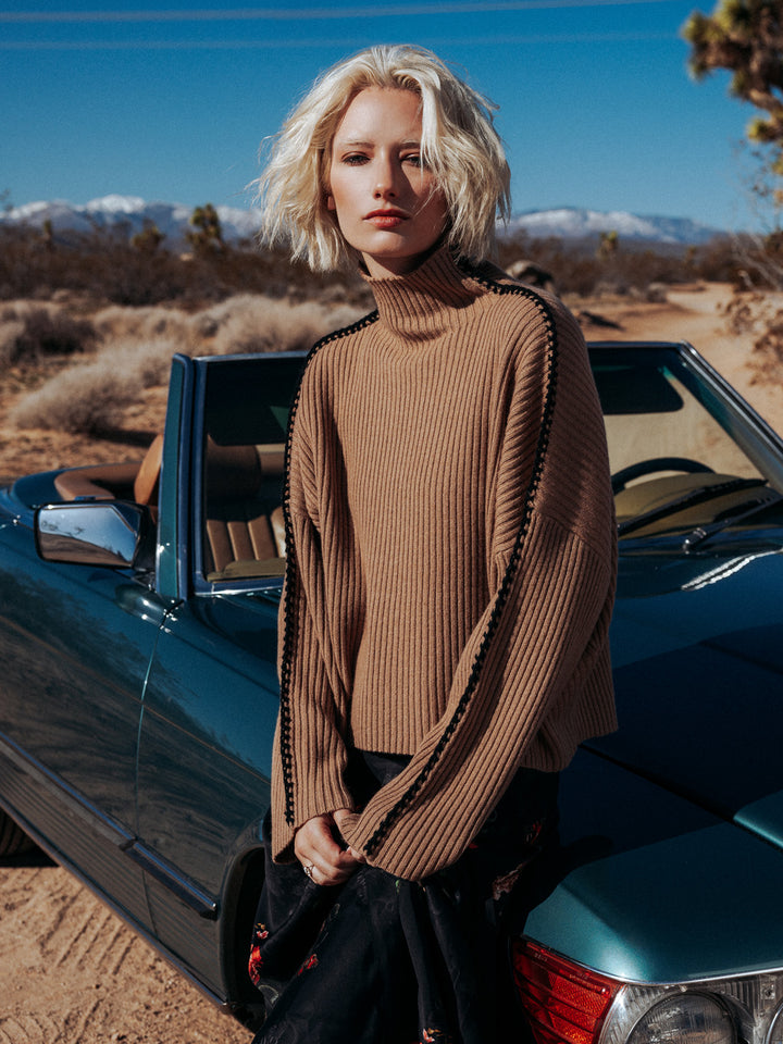 Moody editorial photo of model wearing marullo in camel while sitting on a dark teal car in the desert.