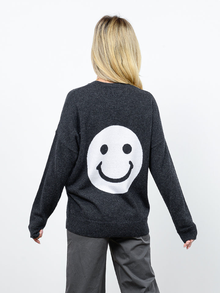 3/4 back view of model wearing Jezebel in gravel grey. The sweater is oversized and made of 100% cashmere. The crewneck pullover has a large smiley face graphic on the back.