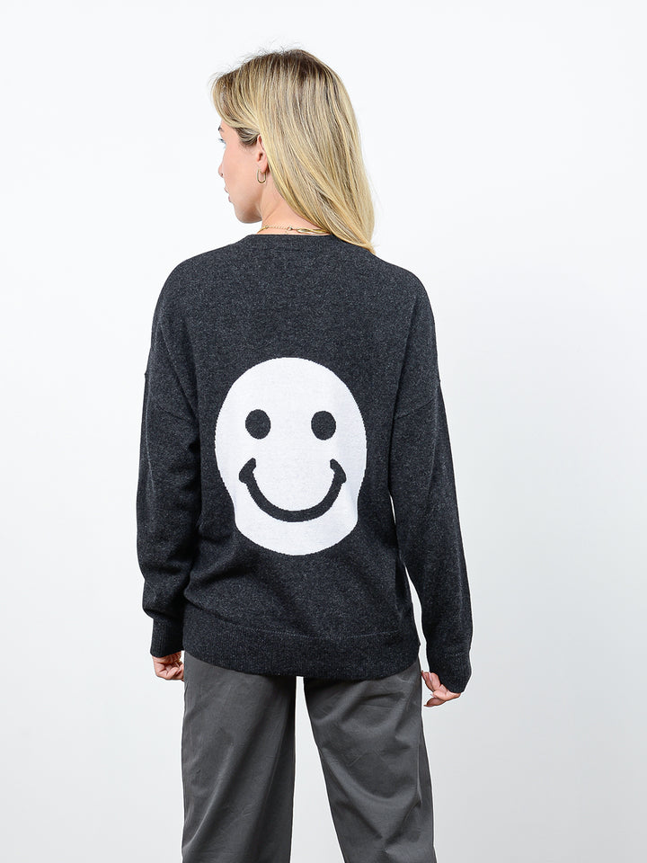 Back facing shot of model wearing Jezebel in gravel grey. The sweater is oversized and made of 100% cashmere. The crewneck pullover has a large smiley face graphic on the back.