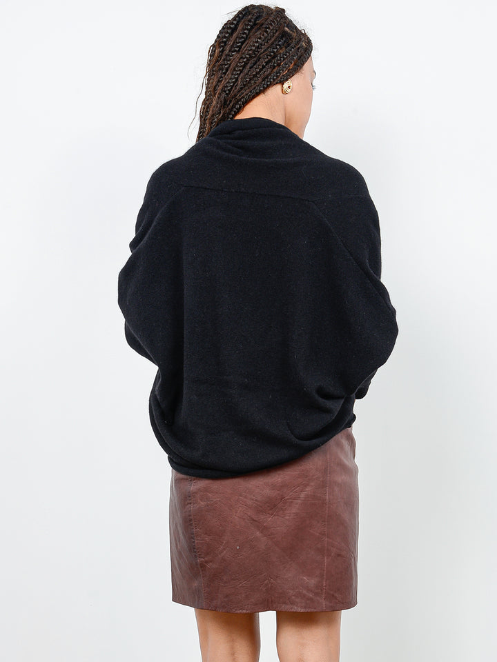 Back facing shot of model wearing Kirra in black. The sweater is oversized and made of cashmere and recycled cashmere. The sweater has a draped cowl neck with dolman sleeves that are fitted at the forearm.