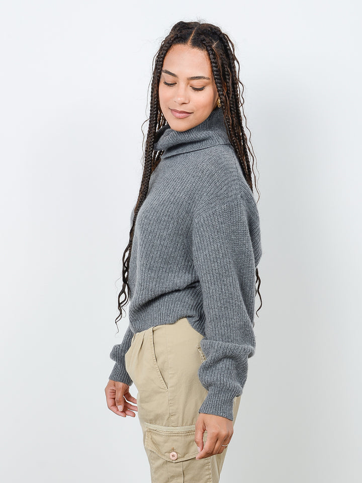 Side view shot of model wearing Naia in gravel gray. The sweater is oversized and cropped and made of cashmere and recycled cashmere. The sweater is a ribbed knit turtleneck.